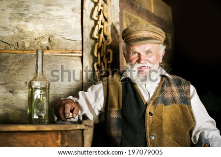 Portrait of old farmer with beard and hat holding a bottle of herbal spirit