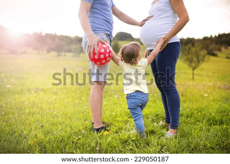 Happy pregnant family with little girl having fun playing with ball in summer nature