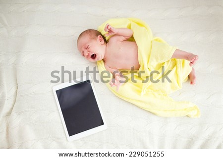 Crying newborn baby girl lying on bed next to digital tablet