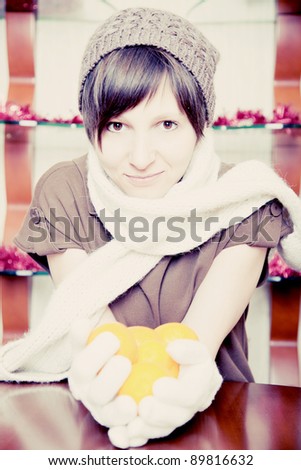 Young adult woman sitting at a table in winter gloves, scarf and hat. holds in his hands tangerines  toned image