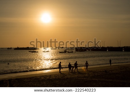 silhouettes of people walking on the beach at sunset, with fishing boats near a jetty.