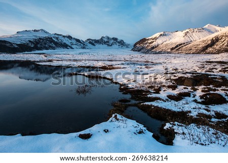 The scenic Vatnajokull National Park in the late afternoon, with snow covered mountains and volcanoes, and calm lakes reflecting the blue skies.
