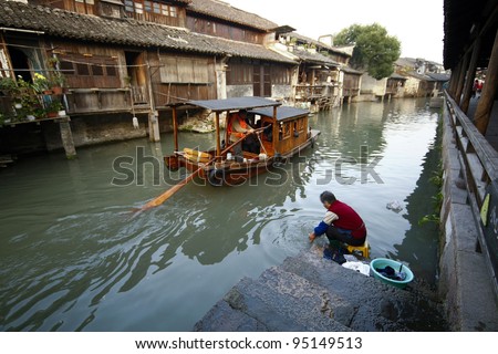 WUZHEN, CHINA - NOVEMBER 25: Tourists in a paddle boat watch a local lady wash her clothes the traditional way in the river in this 1300 years old water town on November 25, 2011 in Wuzhen, China.