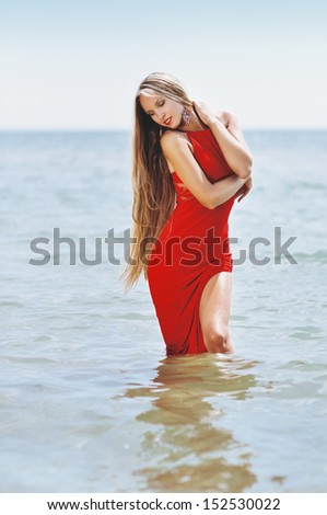 the beautiful woman with smart long hair in a long evening red dress in water on the seashore