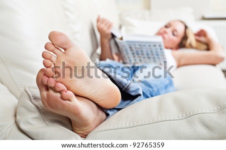 Barefoot young woman lying on sofa and reading book, shallow depth of field, focus on foot soles