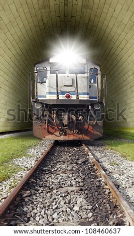 A vintage diesel powered train with a lighted head lamp emerging from a railway tunnel.