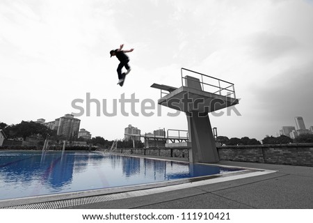 Silhouette of an energetic skateboarder jumping off a diving platform against a dramatic cloudy sky into a blue swimming pool.