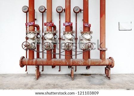 A row of red color fire fighting water supply pipeline system with valves and alarm bell hanging on a white concrete wall.