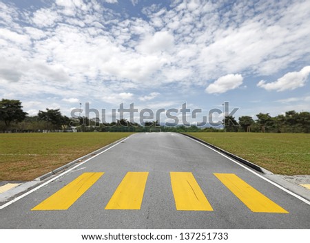 An asphalt road with a yellow zebra crossing on a soccer field leading to the goal mouth on a blue cloudy day.