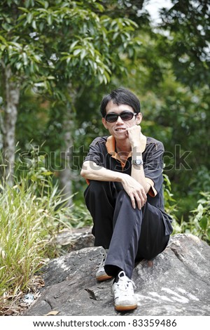 A thin middle aged chinese man wearing a sunglasses sitting on a granite rock in a green outdoor forest park while thinking about the environment.