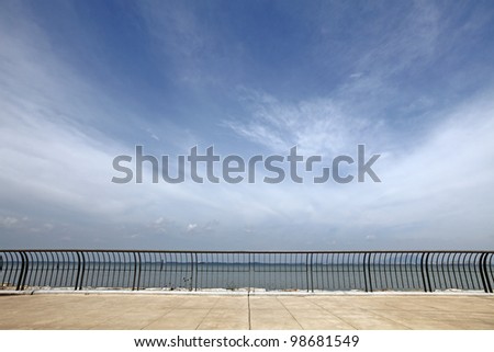 A concrete platform with metal railings overlooking a coastal seafront with dramatic swirling cloud on a blue sky.