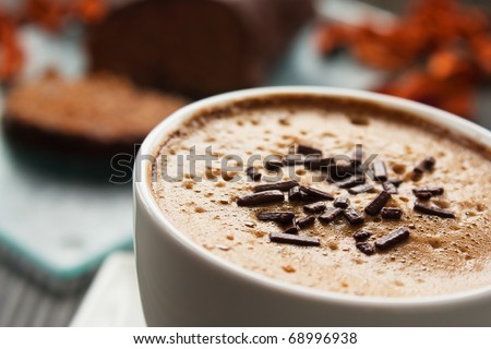 Capuccino in white cup  with chocolate sprinkles and cake in the back.