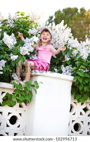 Blond baby girl sitting on fence at lilac, singing song and swinging arms