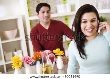 A young woman is talking on a cell phone while a young man stands behind a kitchen counter in the background. Horizontal shot.