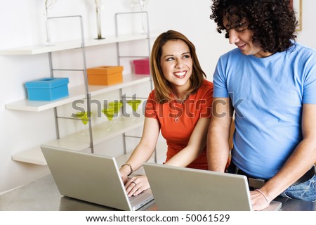 A young man and woman are smiling at one another while working side by side on laptops. Horizontal shot.