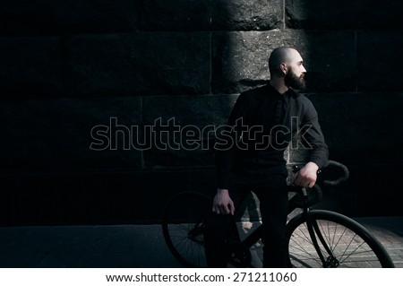 bearded caucasian bald man sits on black fix bicycle against black wall of stone. He sits in thin stripe of light. Street is empty. Man is wearing black shirt and trousers and sneakers.