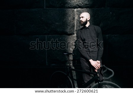 bearded caucasian bald man with black fix bicycle against black wall of stone. He is in thin stripe of light. Street is empty. Man is wearing black shirt and trousers and sneakers.