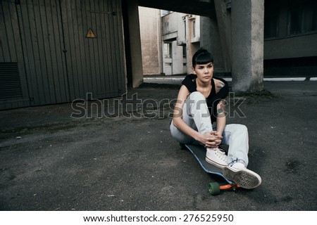 young fit woman with modern haircut sitting on longboard. she is in the shade of building. she wears jeans and singlet. She has tattoos throughout her body. longboard has no prints or aerography