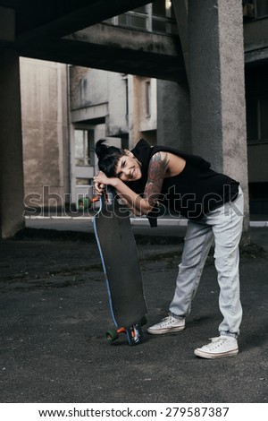 young fit woman with modern haircut standing with longboard. she is in the shade of building. she wears jeans and singlet. She has tattoos throughout her body. longboard has no prints or aerography