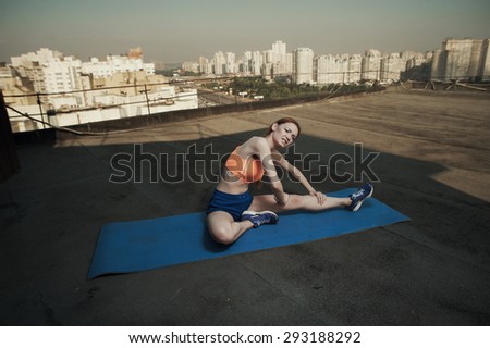 sexy red haired young woman writhing in pain after stretching on mat on high rise roof. she wears blue shorts and orange top and blue sneakers. she is in shade of outbuilding against other high rises