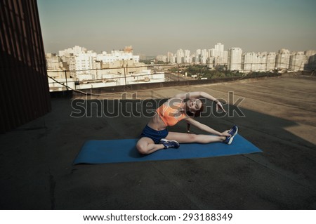 smiling beautiful red haired young lady does stretching on mat on roof of high rise. she wears blue shorts and orange top and blue sneakers. she trains in shade of outbuilding against other high rises