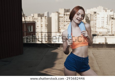 smiling exhausted red haired lady with towel around her neck after workout on roof of high-rise. She wears orange top and blue shorts. Towel is blue. She stands against other high rises