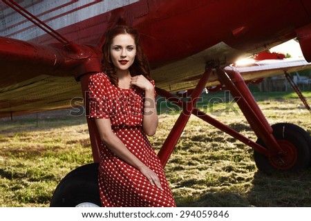 beautiful cheerful brown haired pin-up lady with vintage haircut and red dress sits on airplane wheel in setting sun. The airplane is red and vintage and stands on sunlit grass.