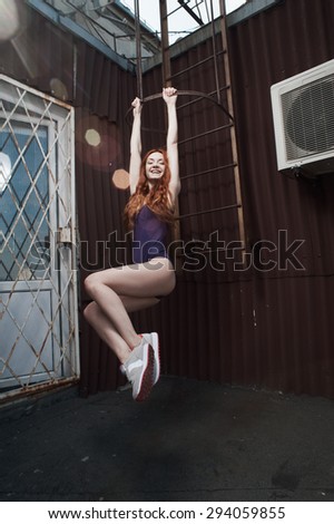 smiling lady with red hair hanging on fire escape on roof of high-rise in purple swimsuit and gray sneakers. she is young and slim. hair fluttering in the wind.