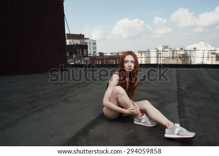 lady with red hair poses on roof of high-rise in purple swimsuit and gray sneakers. she is young and slim. hair fluttering in the wind. she sits against other high-rises and cloudy sky