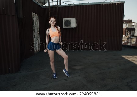 exhausted red haired lady with skipping rope at workout on roof of high-rise. She wears orange top and blue shorts. skipping rope is black. She poses against outbuilding and fire escape. She is slim