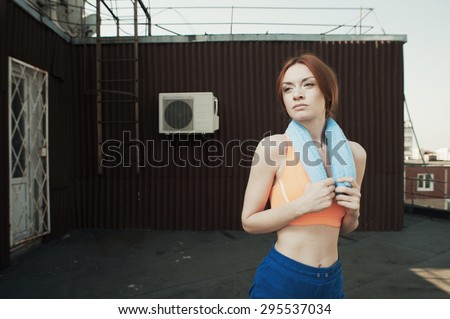 exhausted red haired lady with towel around her neck after workout on roof of high-rise. She wears orange top and blue shorts. Towel is blue. She stands against outbuilding and fire escape.
