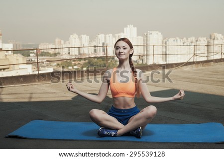 smiling beautiful red haired young woman meditates on mat on roof of high rise. she wears blue shorts and orange top and blue sneakers. she trains in the shade of outbuilding against other high rises