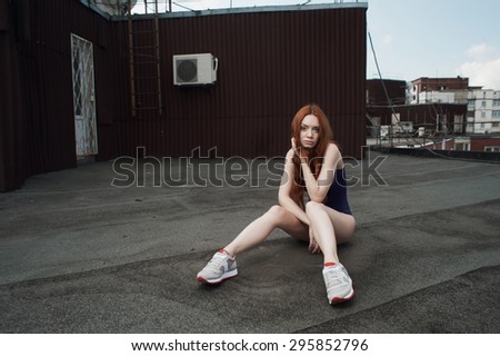 lady with red hair poses on roof of high-rise in purple swimsuit and gray sneakers. she is young and slim. hair fluttering in the wind. she sits against other high-rises and cloudy sky