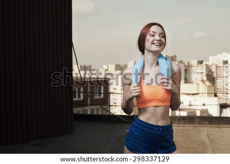 smiling exhausted red haired lady with towel around her neck after workout on roof of high-rise. She wears orange top and blue shorts. She stands against building and other high rises and cloudy sky.