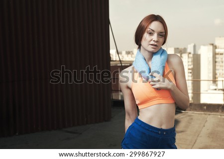 exhausted red haired lady with towel around her neck after workout on roof of high-rise. She wears orange top and blue shorts. She stands against building and other high rises and cloudy sky.