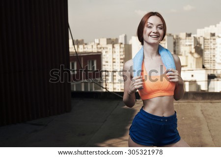 smiling exhausted red haired lady with towel around her neck after workout on roof of high-rise. She wears orange top and blue shorts. She stands against building and other high rises and cloudy sky.