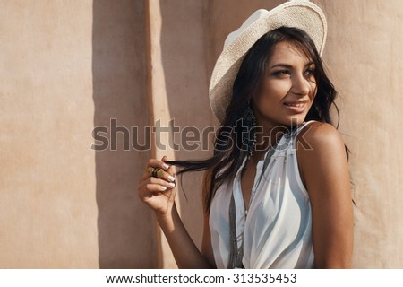 smiling indian lady in jeans, white shirt and white hat against ancient building. She is in harsh morning light. She is positive and playful.