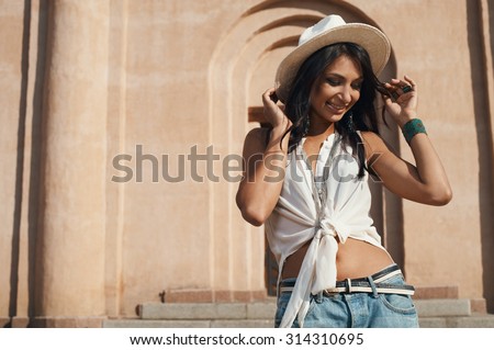 smiling indian lady in jeans, white shirt and white hat against ancient building. She is in harsh morning light. She is positive and playful. Building looks like church or eastern temple