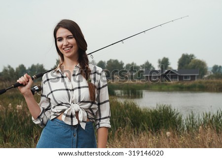 Cute rural brown haired lady posing against ranch house and pond with fishing rod. She stands in grass against rural scape. She wears jeans dress. House is made of wood and is brown. Fish-rod is black