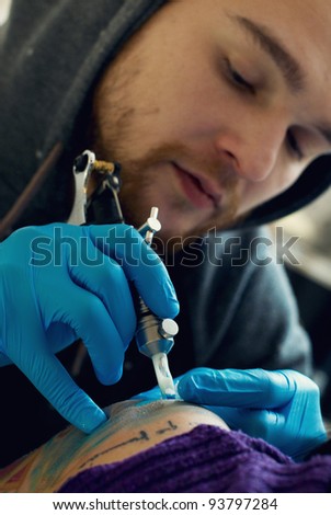 Bearded Caucasian tattooist creates a tattoo on a woman\'s shoulder, close up. He is wearing blue gloves