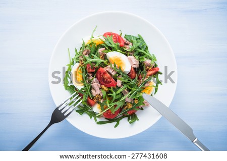 Fresh salad with tuna, tomatoes, eggs, arugula and mustard on blue wooden background top view. Healthy food.