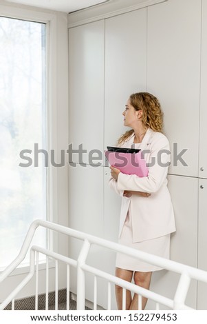 Serious thoughtful business woman standing in office hallway holding binder looking out window