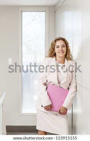 Smiling successful business woman standing in office hallway holding binder