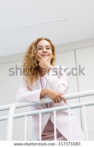 Portrait of confident successful business woman standing in office hallway leaning on railing shot from below