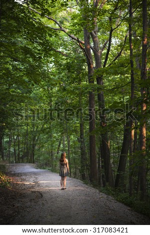 Small figure of young woman standing on path in dark forest with big tall trees illuminated by evening sunshine