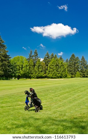 A golf bag on three wheel golf cart over a beautiful green with blue sky and white clouds.
