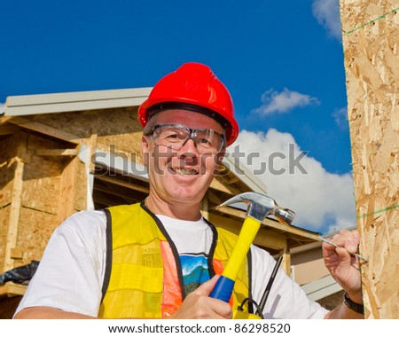 A man in a hard hat standing in front of an house holding a hammer and nail.