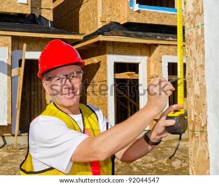 A man in a hard hat standing in front of an house at sunny day holding a measure tape in his hand.