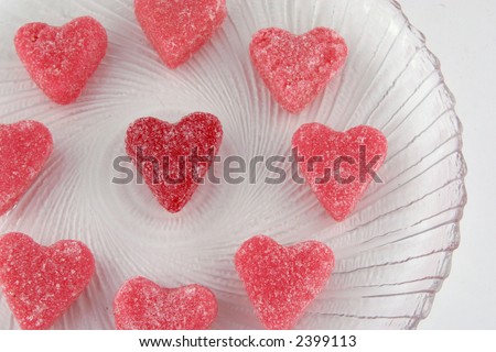 Pink heart shaped candies surrounding a red one, all sugar coated, on a clear glass dish.
