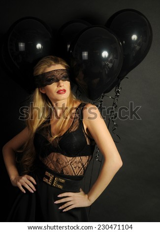 Young beautiful slim girl wearing black lace sensual top and black skirt with evening make up and black lace band on her eyes posing with black balloons on black background
